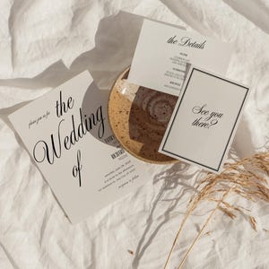 May include: Wedding stationery set in a beige palette. The invitation cards display elegant black script, including the phrases "the Wedding of" and "See you there?" A ceramic plate and dried grass complete the arrangement.