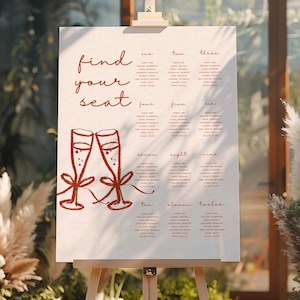 Pode incluir: Um grande quadro de assentos de casamento branco sujo em um cavalete. O gráfico apresenta uma escrita vermelha que diz "find your seat" e um par de taças de champanhe. Os nomes dos convidados estão listados sob os números de um a doze.