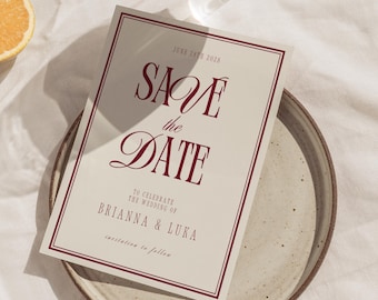 May include: A rectangular wedding "Save the Date" card with burgundy text and border. The card reads "SAVE the DATE" and "TO CELEBRATE THE WEDDING OF BRIANNA & LUKA". The card is on a neutral-toned plate.