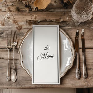 Może przedstawiać: Formalne nakrycie stołu z białym talerzem, sztućcami i kartą menu z napisem "the Menu". Karta menu jest wyśrodkowana na talerzu, z nożami i widelcami po obu stronach. Stół jest drewniany.