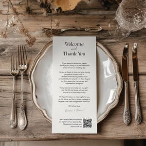 Può includere: Un'apparecchiatura per la tavola nuziale con un piatto bianco, posate e un biglietto di benvenuto. Il biglietto dice "Benvenuti e Grazie" e include un codice QR. L'allestimento è su una superficie di legno.