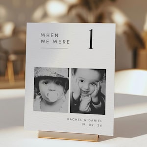 May include: A black and white photo card with the text "WHEN WE WERE" and the number "1". The card features two baby photos and the names "RACHEL & DANIEL" with the date "19. 02. 26". The card is displayed in a gold-colored holder.