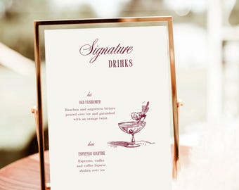 Puede incluir: Un letrero con marco dorado con las palabras "Signature Drinks" en escritura burdeos. El letrero enumera dos recetas de cócteles: "Old Fashioned" y "Espresso Martini", cada una con una breve descripción y una ilustración. El letrero está sobre una superficie de madera.