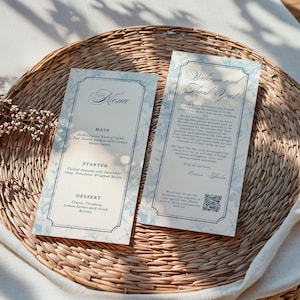 Może przedstawiać: Dwie eleganckie karty menu weselnego z kwiatowym wzorem w kolorze niebieskim i białym. Menu zawiera dania główne, przystawki i desery. Druga karta zawiera napis "Witamy i dziękujemy!" z kodem QR.