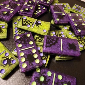 May include: A set of Halloween-themed dominoes with a spider web and spider design. The dominoes are made of resin and have a glittery finish. The dominoes are green and purple.