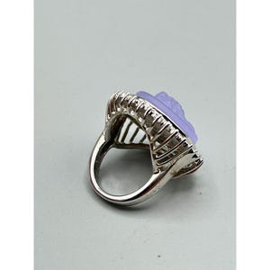 May include: A silver ring featuring a light purple, carved floral design. The ring has a rectangular setting with a detailed, textured border. The band is smooth and polished, and the ring is set against a light gray background.