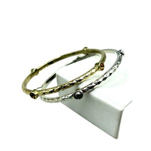 May include: Three metal bangles in gold and silver tones. The gold bangle has a textured surface and is adorned with small, round, brown gemstones. The silver bangle has a similar texture and features a dark gray gemstone. The bangles are displayed on a white surface.