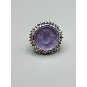 May include: A silver-toned ring featuring a round, lavender-colored stone with a carved face. The stone is surrounded by a halo of iridescent, multi-colored crystals. The ring has a detailed, ornate design, suitable for jewelry collections.