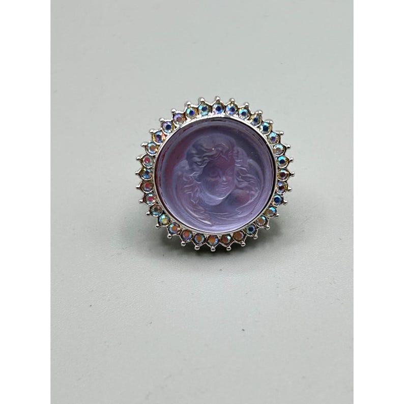 May include: A silver-toned ring featuring a round, lavender-colored stone with a carved face. The stone is surrounded by a halo of iridescent, multi-colored crystals. The ring has a detailed, ornate design, suitable for jewelry collections.