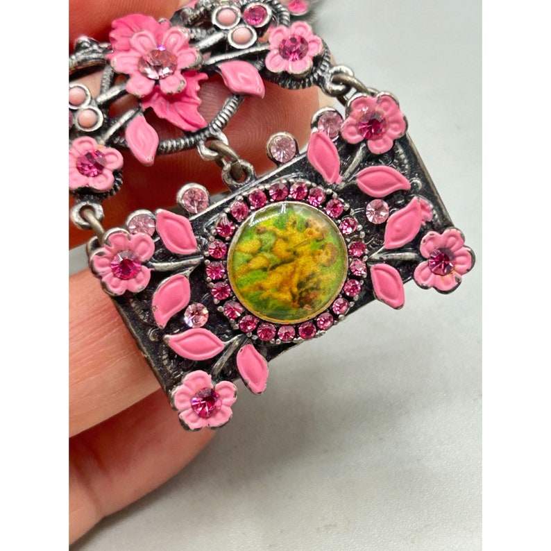 May include: A pink and silver pendant necklace with a floral design and a small round cameo with a green and yellow image. The pendant is surrounded by pink rhinestones.