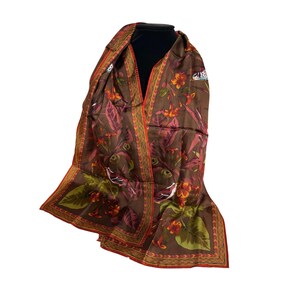 May include: A brown scarf featuring a botanical print with orange and red flowers, green leaves, and small birds. The scarf has a decorative border with a repeating geometric pattern in shades of orange and red along the edges of the fabric.