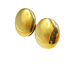 May include: Two gold-colored, oval-shaped clip-on earrings. Each earring has a smooth, reflective surface with a curved, recessed detail near the bottom. The earrings are set against a white background, highlighting their metallic sheen.