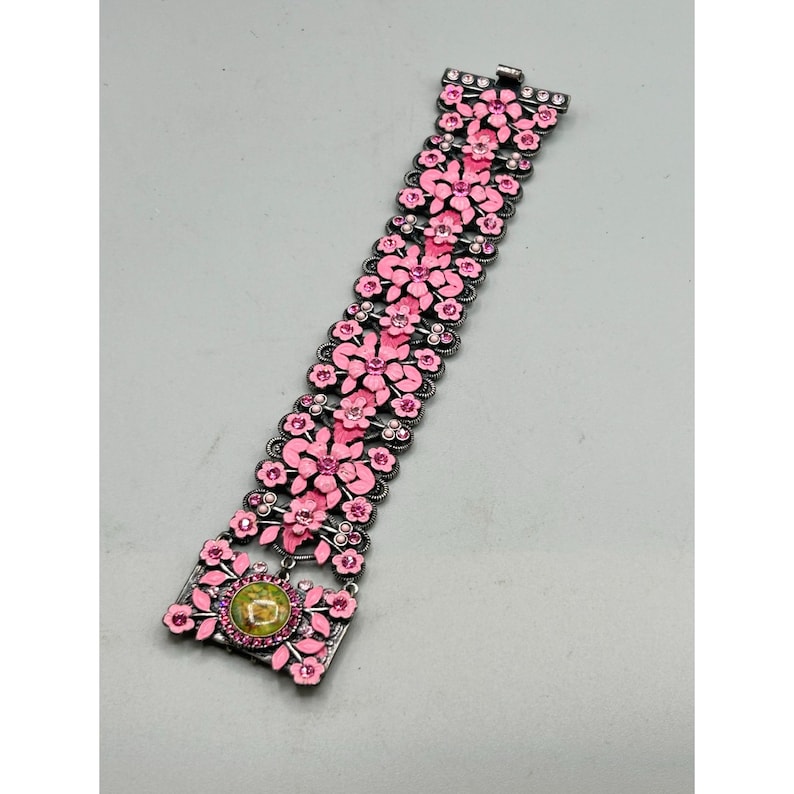 May include: A pink and silver floral bracelet with a green stone in the center. The bracelet is made of metal and has a delicate design.