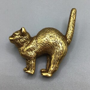 Puede incluir: Una figurita de metal de color dorado de un gato en posición agachada. El gato tiene una superficie detallada y texturizada y una cola larga.