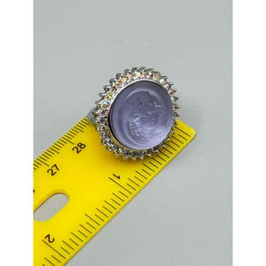 May include: A silver-toned ring with a large, round, lavender-colored stone. The stone features a carved face and is surrounded by a halo of iridescent crystals. The ring is measured with a yellow ruler.