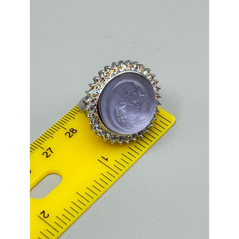 May include: A silver-toned ring with a large, round, lavender-colored stone. The stone features a carved face and is surrounded by a halo of iridescent crystals. The ring is measured with a yellow ruler.