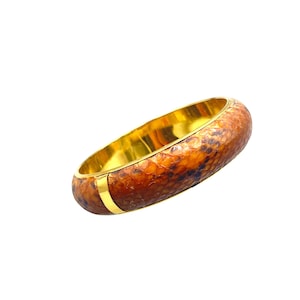 Puede incluir: Un brazalete dorado con un patrón de piel de serpiente texturizado en tonos marrón y naranja. El interior es dorado, con una banda dorada en el exterior. Esta joya es un accesorio llamativo.
