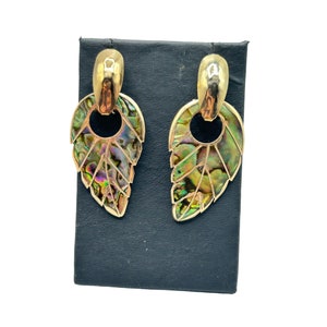 May include: A pair of gold-toned leaf earrings with iridescent abalone shell inlays. The earrings have a post-back closure and a unique, nature-inspired design. The earrings are displayed on a black jewelry stand.