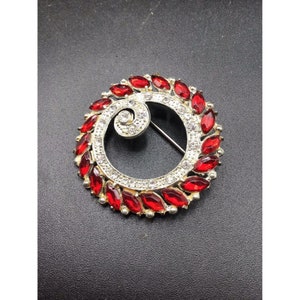 Pode incluir: Um broche de prata com um design circular. O centro do broche é um redemoinho de strass transparentes. A borda externa do broche é um círculo de strass vermelhos.