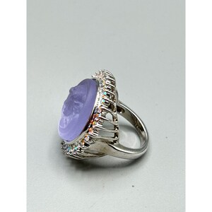 May include: A silver-toned ring featuring a large, oval, lavender-colored stone with a carved profile. The stone is surrounded by a halo of small, iridescent crystals. The ring's band is a simple, polished silver.