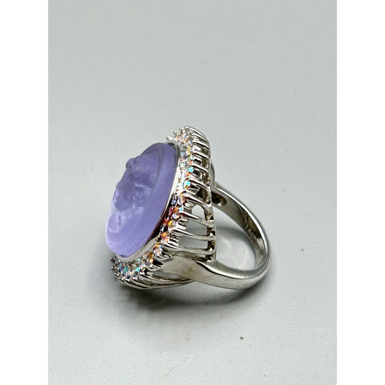 May include: A silver-toned ring featuring a large, oval, lavender-colored stone with a carved profile. The stone is surrounded by a halo of small, iridescent crystals. The ring's band is a simple, polished silver.