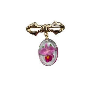 Könnte beinhalten: Eine goldene Schleifenbrosche mit einem klaren ovalen Anhänger, der eine gepresste rosa Orchidee enthält.