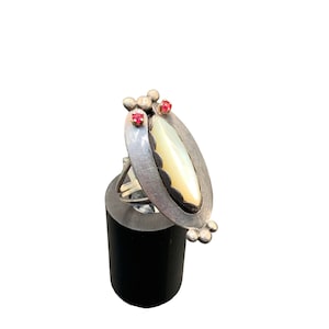 May include: A silver ring with a white oval shaped stone set in a silver frame. The ring has two small red stones on the top.