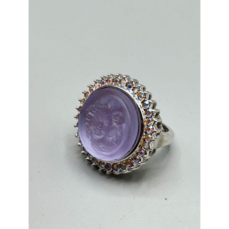 May include: A silver ring featuring a large, oval, lavender-colored stone with a carved profile of a woman. The stone is surrounded by a halo of small, iridescent gemstones. The ring has a wide band.