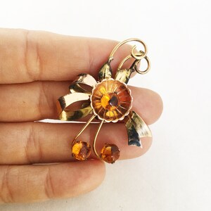 May include: A gold-toned brooch with a flower design featuring amber-colored glass stones. The flower has a bow-like shape with two smaller amber stones on the bottom.