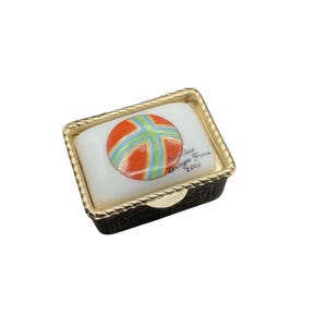 May include: A small rectangular box with a gold trim and a white porcelain lid. The lid features a colorful ball with orange, blue, and green stripes. The text "Clair Limoges France 2000" is printed on the lid.