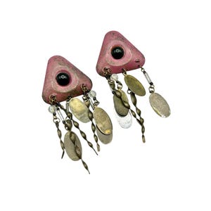May include: A pair of vintage earrings. Each earring features a pink, triangular base with a black center and dangling metal accents. The dangling elements include oval-shaped pieces in silver and gold tones, along with clear beads and twisted metal strands.