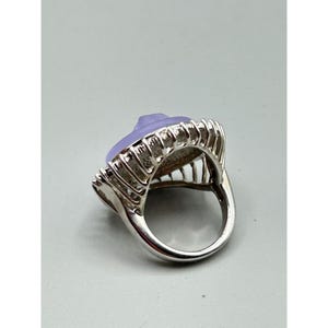 May include: A silver ring with a large, oval, lavender-colored stone. The stone is set in a decorative silver setting with vertical bars. The ring has a wide band and a polished finish. The ring is likely a fashion accessory.