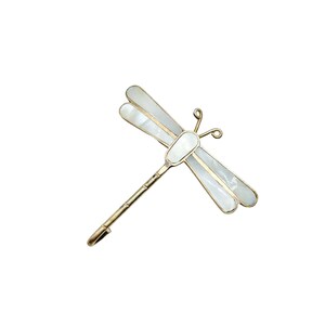 May include: A gold-toned dragonfly hair clip with white iridescent wings. The dragonfly's body and wings are outlined in gold, with a delicate, curved antenna design. The clip is designed to secure hair.