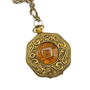 Könnte beinhalten: Eine goldfarbene Halskette mit einem achteckigen Anhänger, der mit einem bernsteinfarbenen Glas-Kamee verziert ist. Der Anhänger ist mit filigranen floralen Mustern verziert.