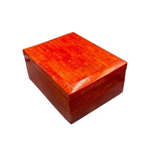 May include: A rectangular wooden box with a reddish-orange finish. The lid and sides have a glossy appearance. The box is made of wood and has a rectangular shape. The box is likely used for storage or display.