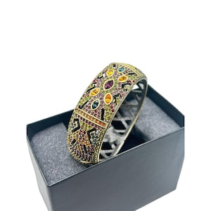 Könnte beinhalten: Ein goldfarbenes Gelenkarmband mit einer farbenfrohen Auswahl an Edelsteinen. Das Armband weist ein geometrisches Design mit einer Vielzahl von Farben auf, darunter Lila, Gelb, Orange und Grün. Es wird in einer schwarzen Box präsentiert.