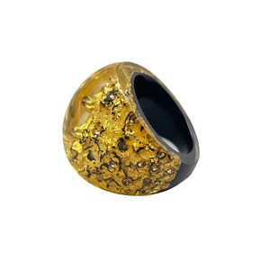 Puede incluir: Un anillo negro con una cúpula de resina transparente y dorada con un diseño dorado texturizado.