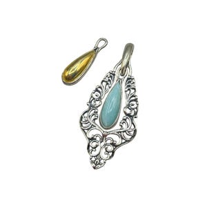 Könnte beinhalten: Zwei Anhänger auf weißem Hintergrund. Einer ist eine silberne Filigran-Tropfenform mit einem hellblauen Stein. Der andere ist ein kleiner, goldfarbener Tropfenanhänger. Beide haben eine Schlaufe für eine Kette.