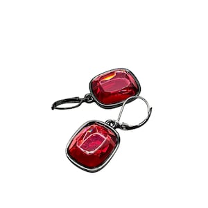 May include: A pair of red gemstone earrings with a square cut. The earrings have a dark metal frame and a lever back closure. The gemstones are a vibrant red color and reflect light.