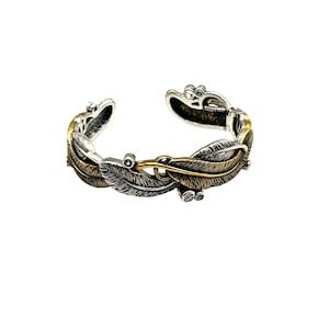 May include: Silver-tone cuff bracelet with a feather design. The bracelet features alternating silver and gold-tone feathers, with small blue accents. The open cuff design allows for easy wear and adjustment. A stylish accessory.