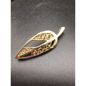 May include: A gold-toned leaf-shaped pendant with a delicate, openwork design featuring small circles. The pendant has a loop at the top for attaching to a chain.