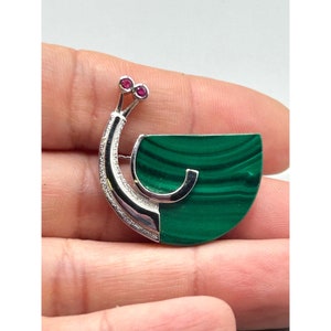 Op de afbeelding: Een zilveren en groene broche met een malachiet-steen en twee robijn-accenten. De broche is gevormd als een gestileerde pauw met een waaiervormige staart.