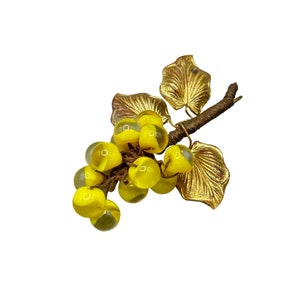Puede incluir: Un broche de tono dorado con forma de vid con uvas de vidrio amarillo. Las hojas son detalladas y las uvas son translúcidas.