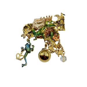 Puede incluir: Un broche de tono dorado con una rana verde que lleva una corona, rodeada de flores, una campana y otros encantos. La rana está posada sobre una hoja de lirio y tiene una corona en la cabeza.