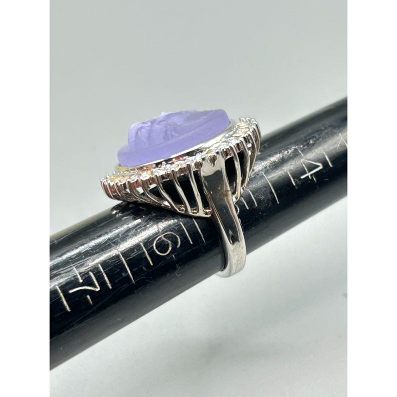 May include: A silver ring with a large, oval, lavender-colored stone. The stone features a carved design and is surrounded by a halo of small, clear stones. The ring is on a black ring sizer.