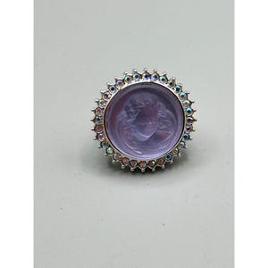 May include: A round, silver-toned ring featuring a lavender-colored center with an engraved angel face. The ring is encircled by a row of iridescent, multi-colored crystals. The ring is set against a light gray background.