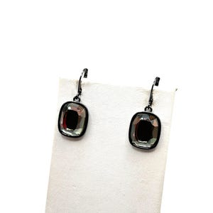 May include: A pair of dangle earrings with dark gray metal frames and large, square-cut, faceted gray gemstones. The earrings are displayed on a white surface against a white background. The gemstones have a reflective quality.