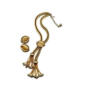 Puede incluir: Un collar de tono dorado con un colgante de perlas y oro y pendientes a juego. El collar tiene un diseño de borla en la parte inferior.