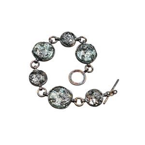 Op de afbeelding: Zilverkleurige armband met zeven ronde, getextureerde bedels met een lichtblauwgroene tint. De bedels zijn verbonden door kleine schakels en decoratieve filigraanelementen. De armband heeft een tuimelsluiting.