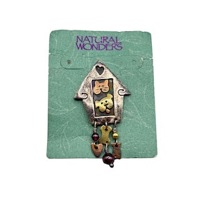 Puede incluir: Un pin de metal plateado en forma de casa con una cara de gato y perro en el interior. El pin tiene un corazón en la parte superior y encantos colgantes que incluyen corazones, huesos y cuentas.